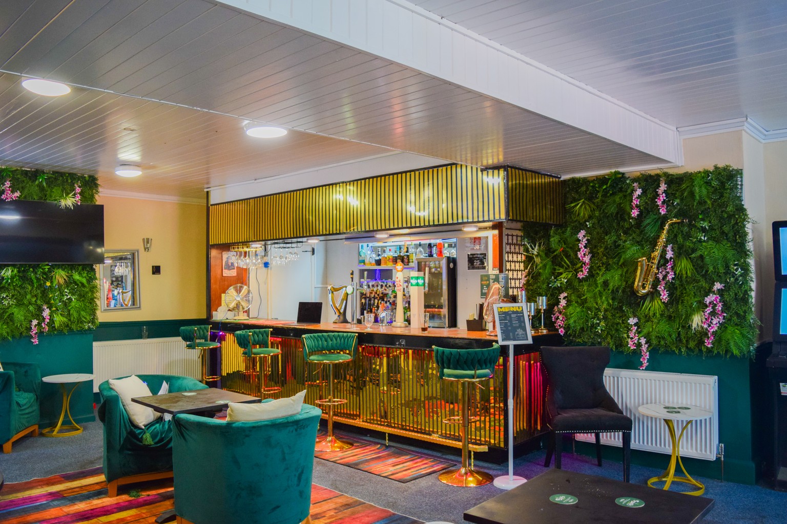 The bar at Ashbury Lounge, with green velvet stools, a foliage wall and lounge seating.